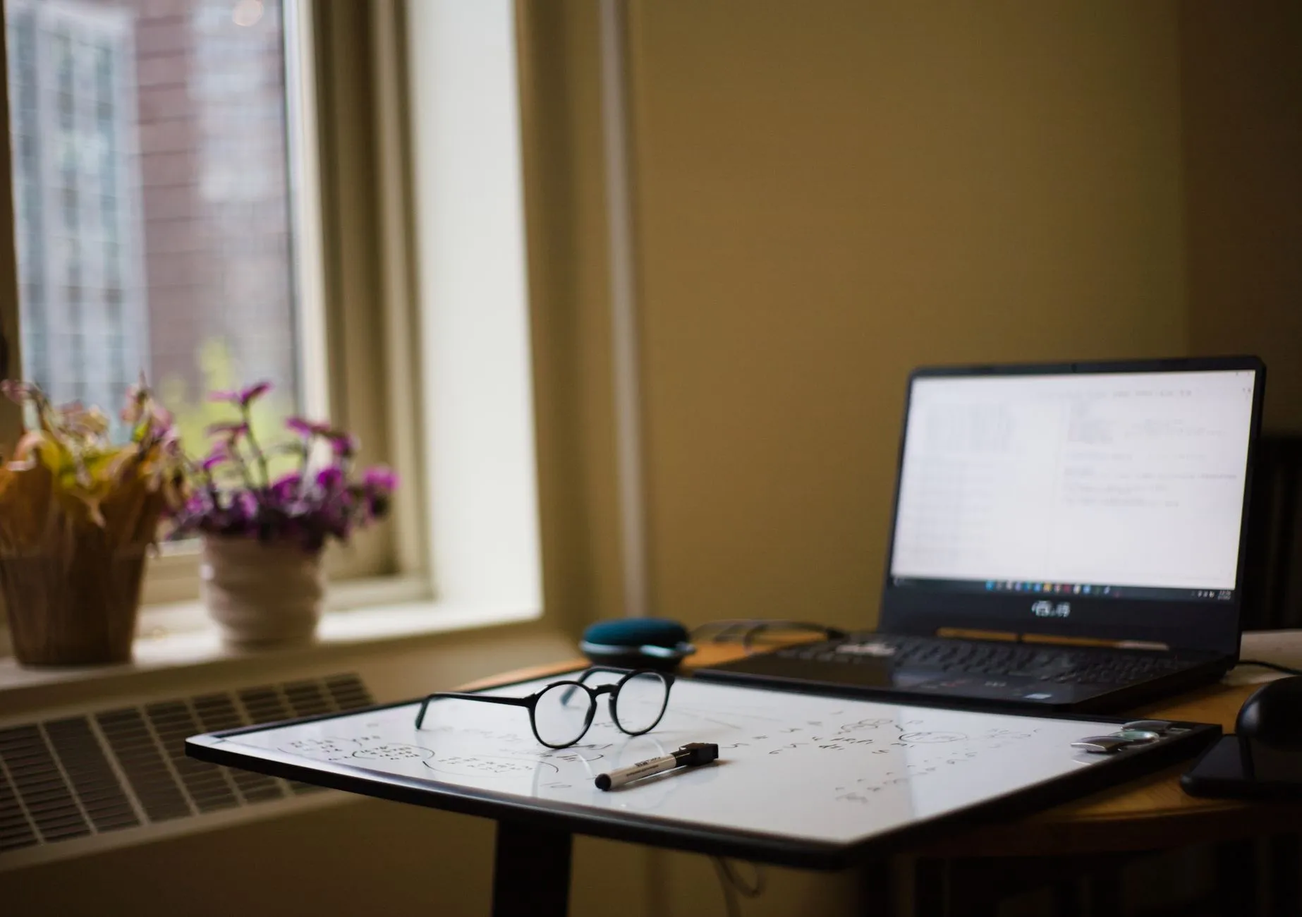 A messy study desk with a laptop and notebooks under soft window light
