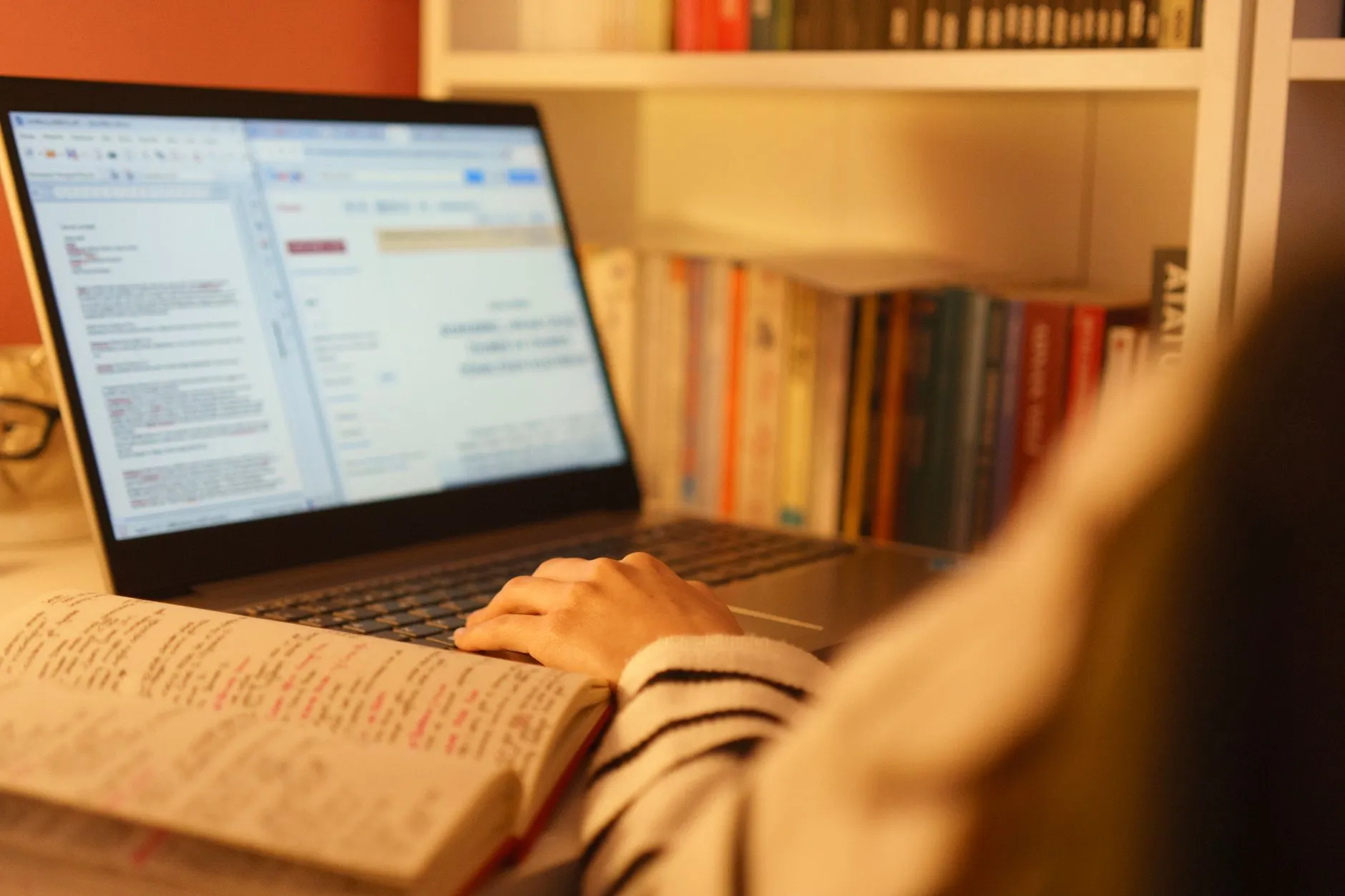 A close-up shot of a student's hands typing on a laptop next to handwritten study notes on a wooden desk