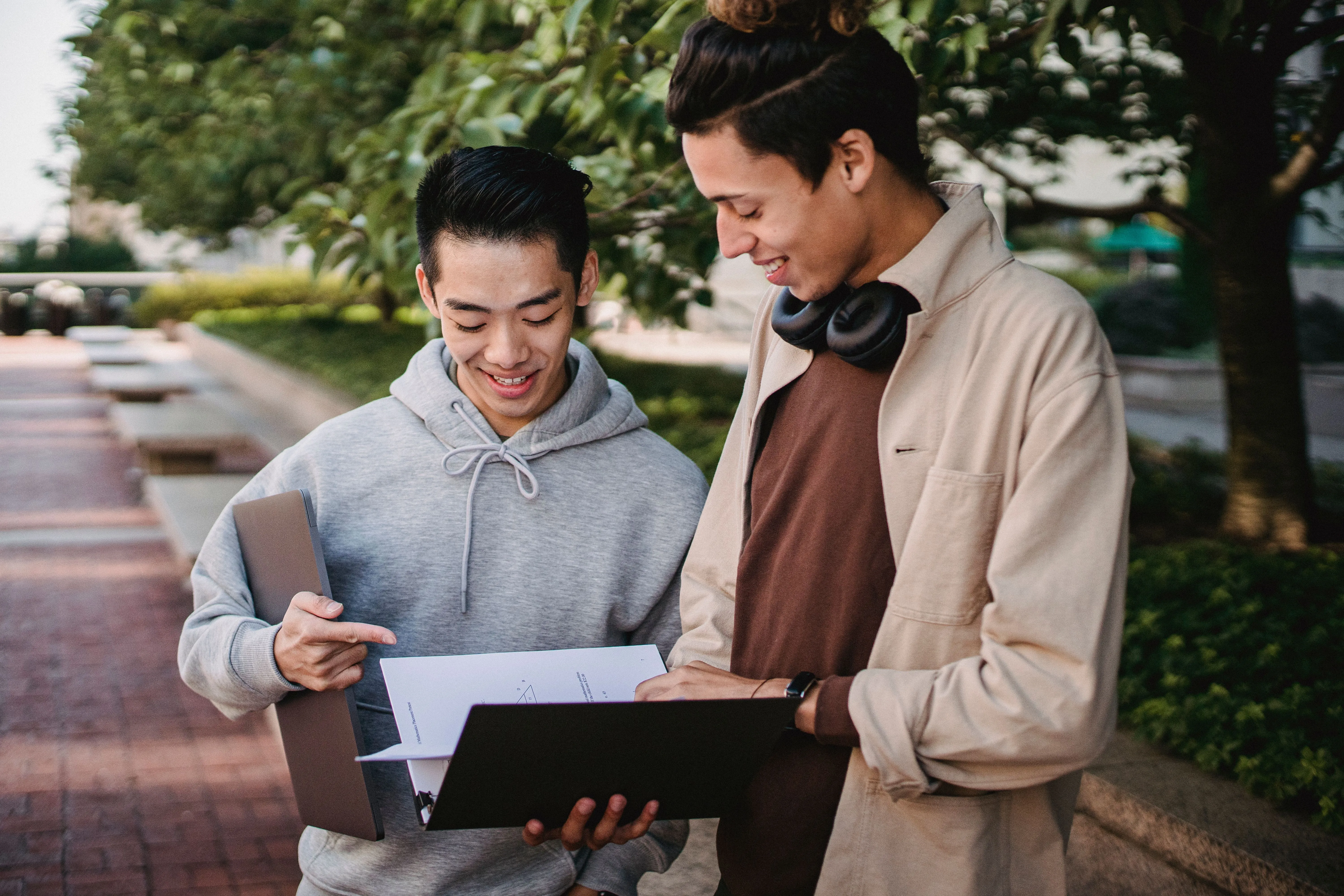 Cheerful multiracial male students in casual clothes looking through pages in folder while standing in green campus park on sunny day