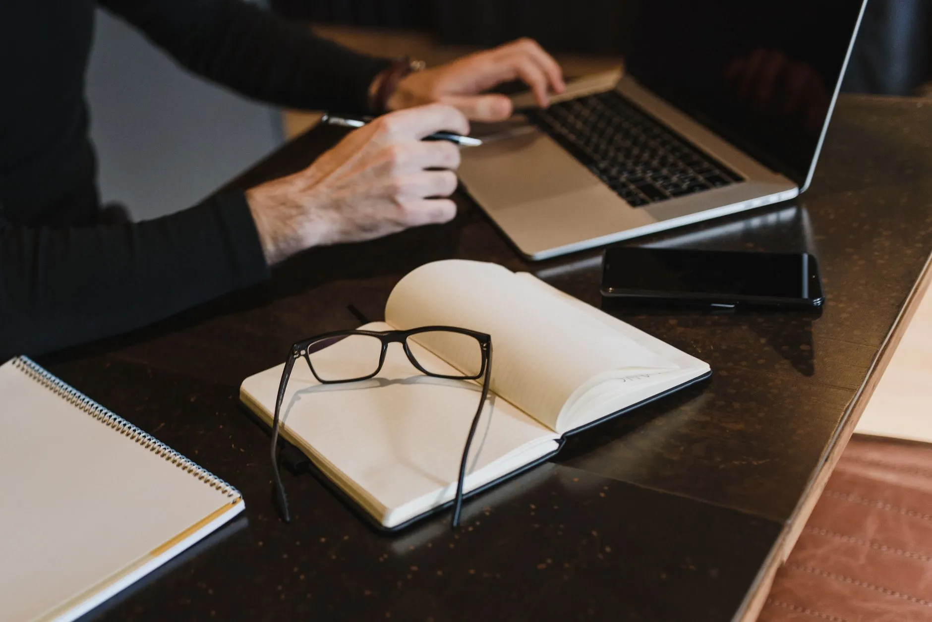 Close up of hands typing on a laptop with a study notebook nearby on a messy desk