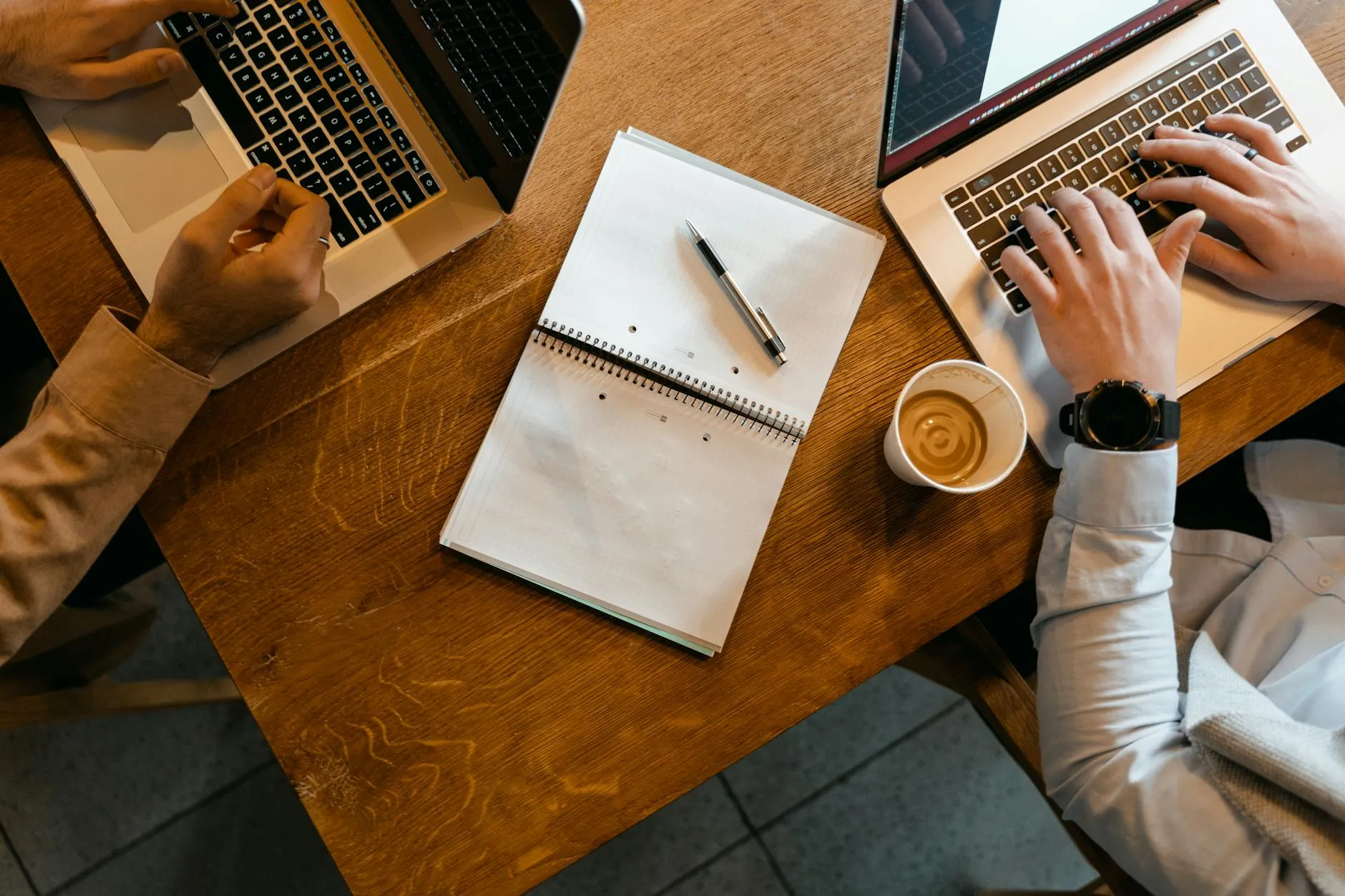 Close up of a student's hands typing on a laptop next to a coffee cup in a busy campus cafe