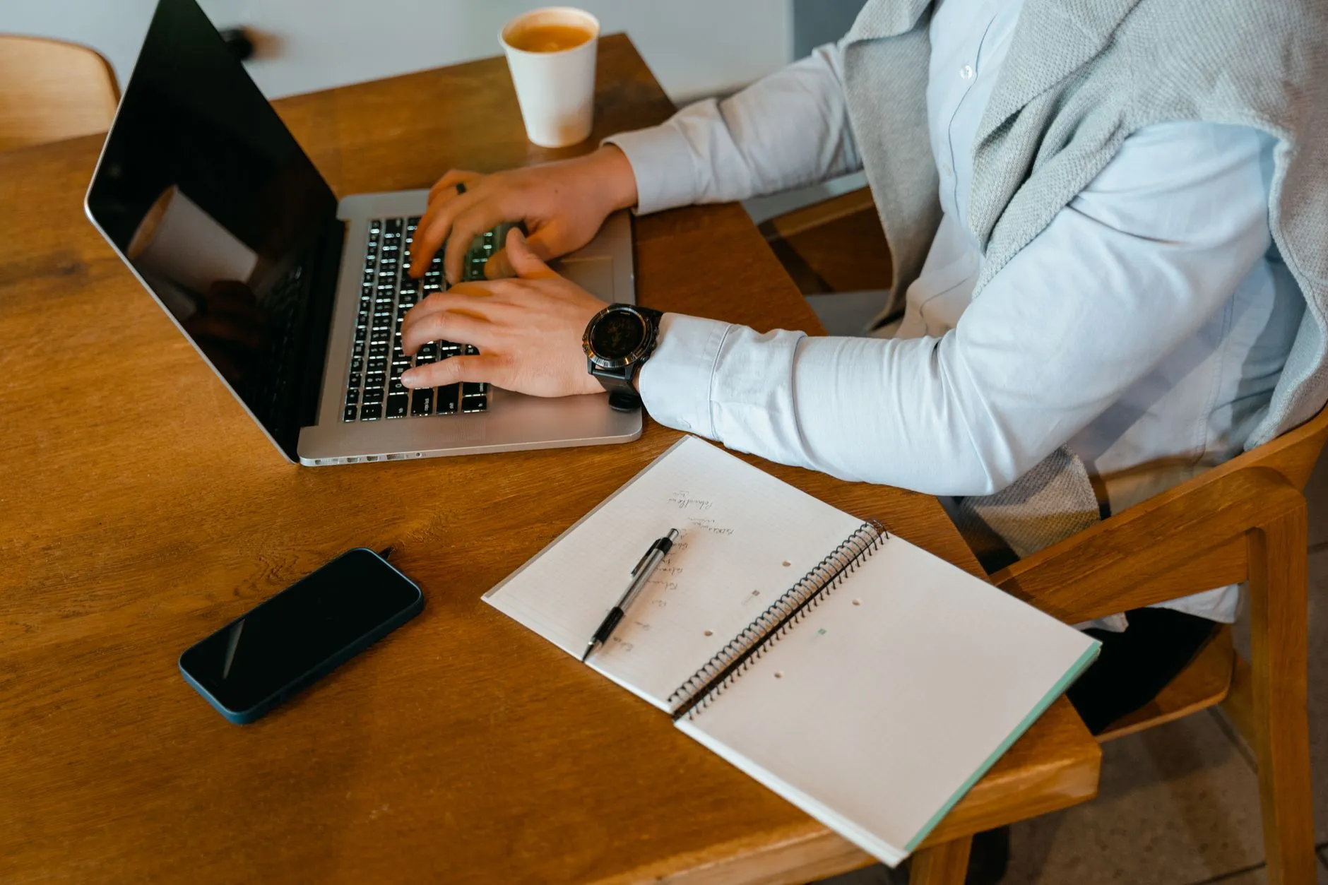 Close up of student hands typing notes on a laptop next to an open textbook on a messy study desk
